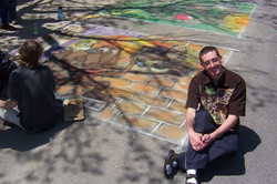 Me sitting next to a mostly finished chalk creation of a dog spray painting a depiction of dogs playing poker